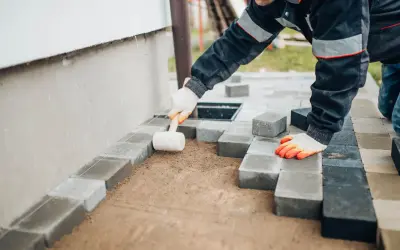 Ręce w rękawiczkach układają kostkę brukową, tworząc opaskę z kostki brukowej przy ścianie domu.
