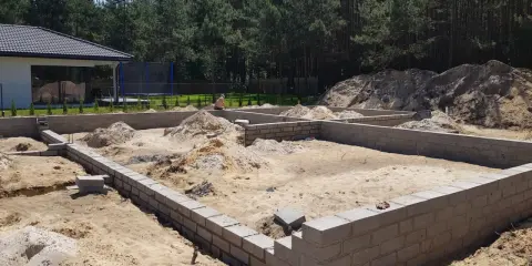 Fundamenty domu w budowie, piasek i stosy ziemi wokół. To etap samodzielnej budowy domu, widać postęp prac.