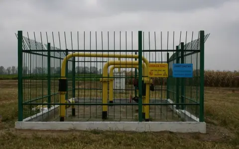 Ogrodzony obiekt gazowy z żółtymi rurami. Tablice ostrzegawcze informują o zagrożeniu. Widać pola uprawne w tle, co sugeruje odległość budynku od gazociągu średniego ciśnienia.