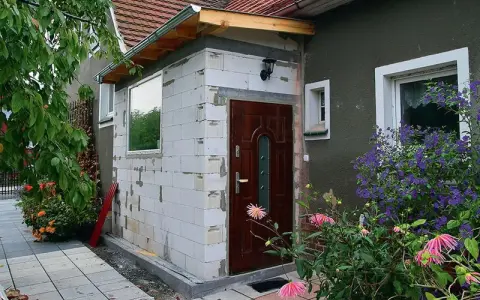 Trwa rozbudowa domu w bok, powstaje nowy wiatrołap z białych pustaków. Obok drzwi wejściowych kwitną kwiaty.