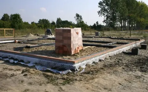 Stan zero budynku: fundamenty z cegły, piasek, folia i stos cegieł pod przykryciem.