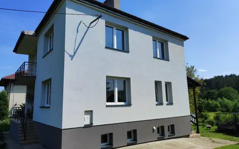Jasny dom typu kostka z szarym cokołem i balkonem, otoczony zielenią.