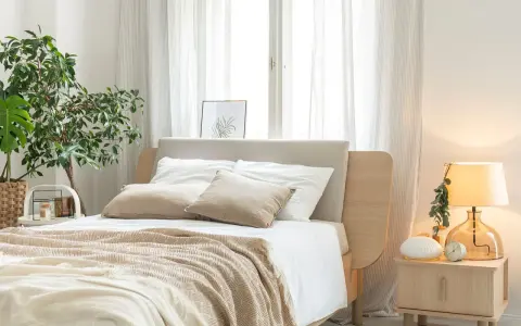 Przytulna sypialnia inspiracje: łóżko z beżowym zagłówkiem, biała pościel, narzuta w jodełkę, lampka nocna i roślina.