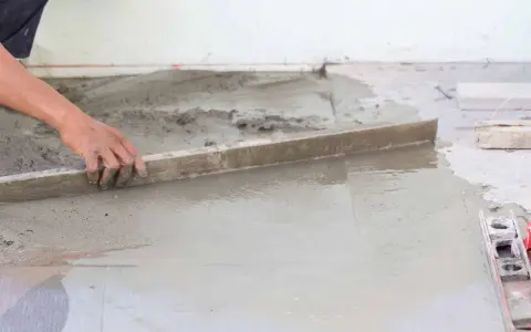 Ręka wyrównuje świeżą betonową wylewkę na ziemi za pomocą łaty. Kluczowe etapy, jak zrobić wylewkę betonową na ziemi.