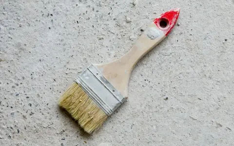 Pędzel do malowania betonu na zewnątrz, z drewnianą rączką i czerwonym zakończeniem, leży na szarej, chropowatej powierzchni.