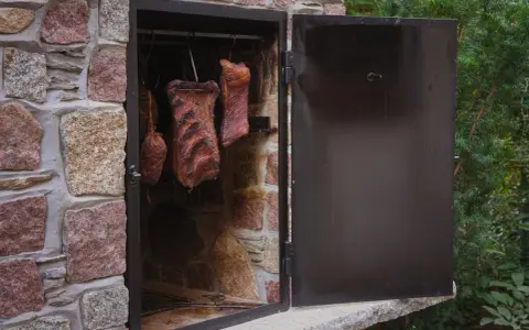 Prosta wędzarnia murowana z wędzonymi mięsami wiszącymi w środku. Drzwi otwarte, widać ruszt.