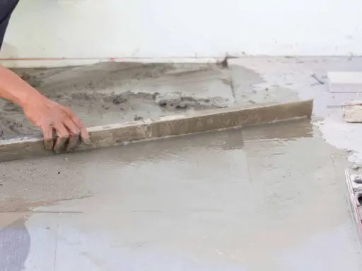 Ręka wyrównuje świeżą betonową wylewkę na ziemi za pomocą łaty. Kluczowe etapy, jak zrobić wylewkę betonową na ziemi.