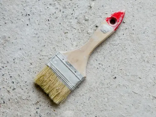Pędzel do malowania betonu na zewnątrz, z drewnianą rączką i czerwonym zakończeniem, leży na szarej, chropowatej powierzchni.