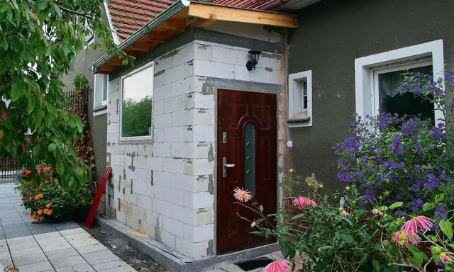 Trwa rozbudowa domu w bok, powstaje nowy wiatrołap z białych pustaków. Obok drzwi wejściowych kwitną kwiaty.