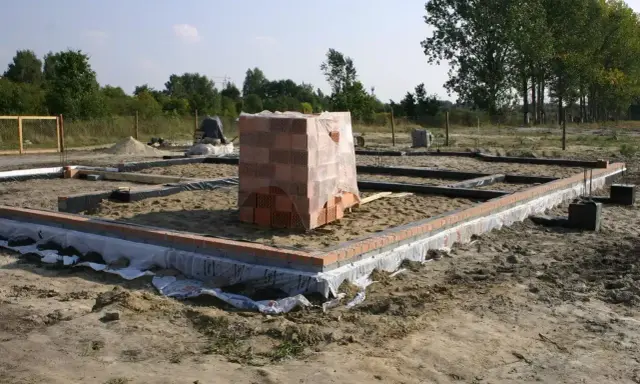 Stan zero budynku: fundamenty z cegły, piasek, folia i stos cegieł pod przykryciem.