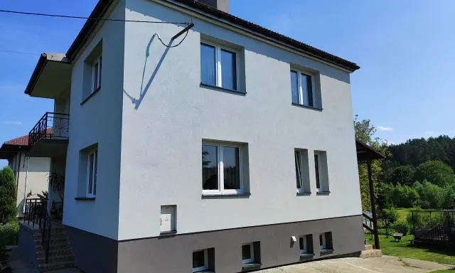 Jasny dom typu kostka z szarym cokołem i balkonem, otoczony zielenią.