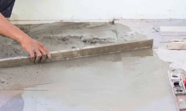 Ręka wyrównuje świeżą betonową wylewkę na ziemi za pomocą łaty. Kluczowe etapy, jak zrobić wylewkę betonową na ziemi.