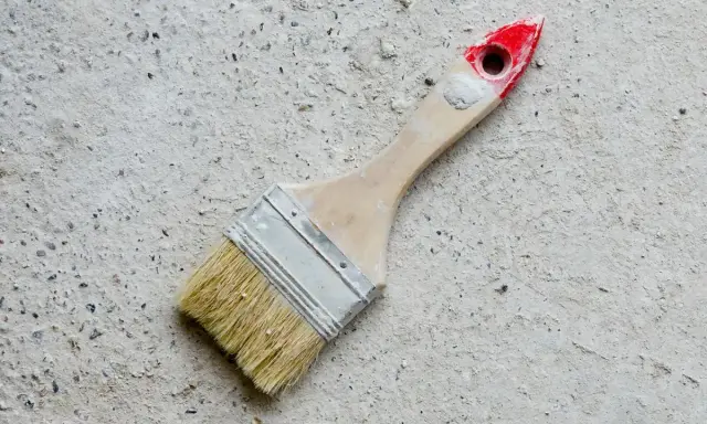 Pędzel do malowania betonu na zewnątrz, z drewnianą rączką i czerwonym zakończeniem, leży na szarej, chropowatej powierzchni.