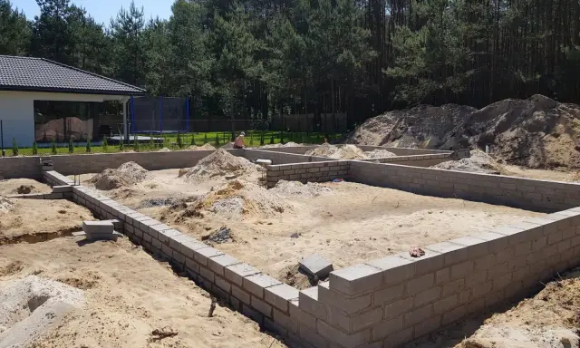 Fundamenty domu w budowie, piasek i stosy ziemi wokół. To etap samodzielnej budowy domu, widać postęp prac.