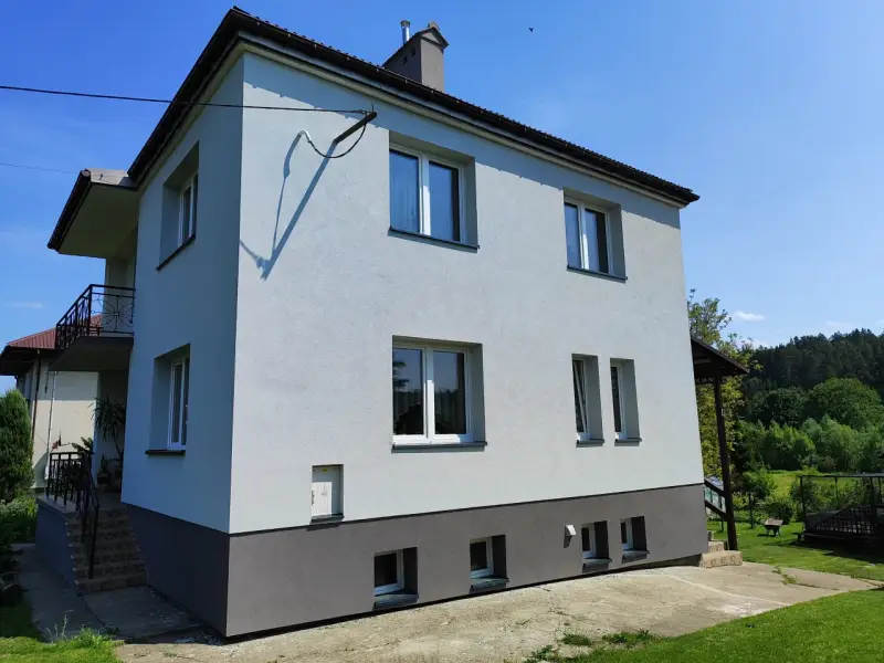 Jasny dom typu kostka z szarym cokołem i balkonem, otoczony zielenią.